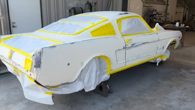 White classic Mustang fastback body shell in the shop, taped off with yellow masking tape, on jack stands.