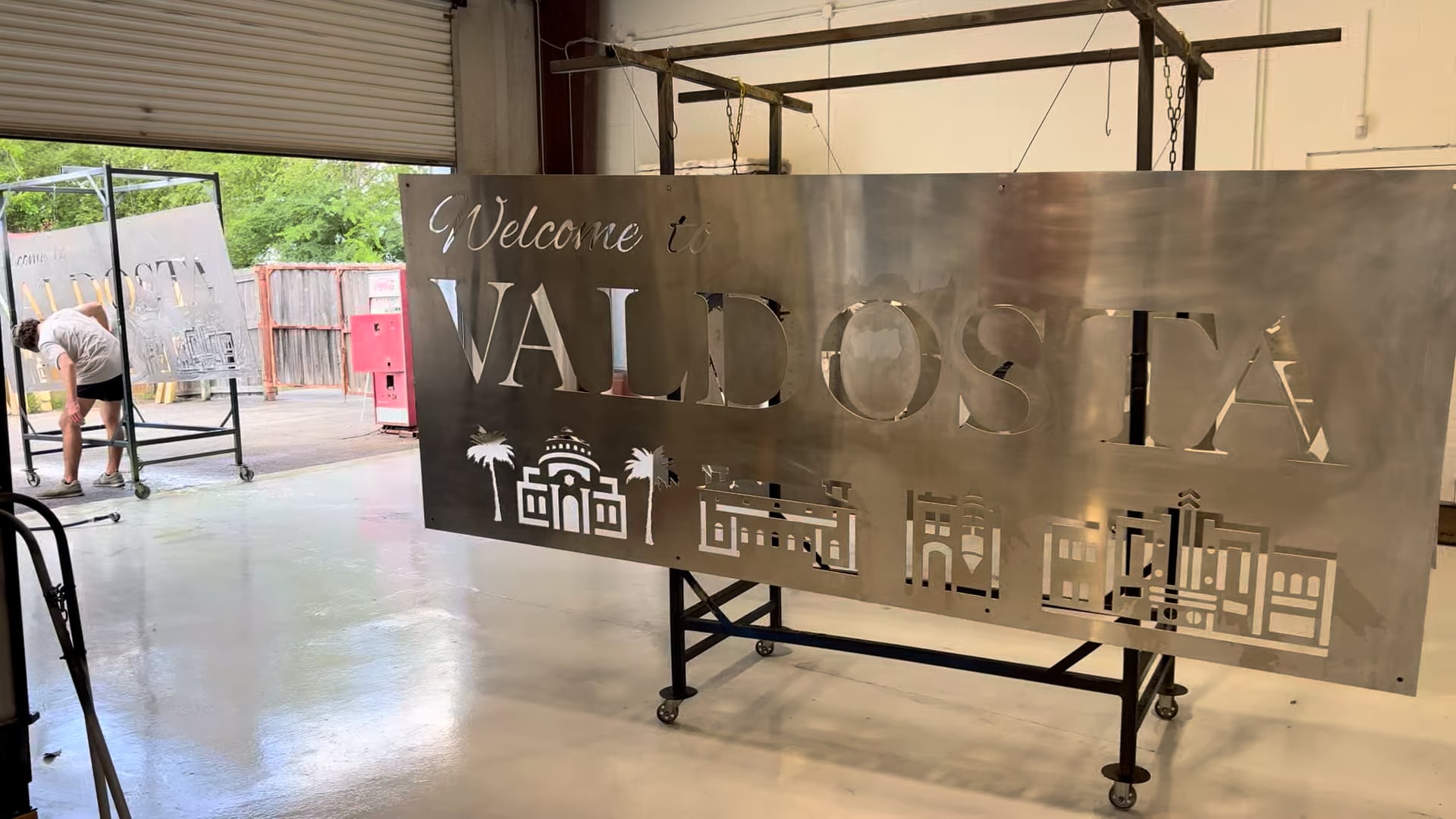 Large cut-metal Welcome to Valdosta sign hanging on a rack inside the shop, roll-up door open behind it.